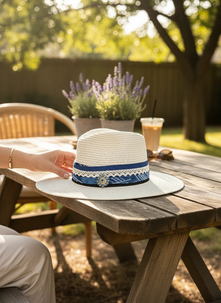 Adams + Hegwood woven straw fedora with a blue patterned fabric band, lace trim, and decorative brooch on a table.