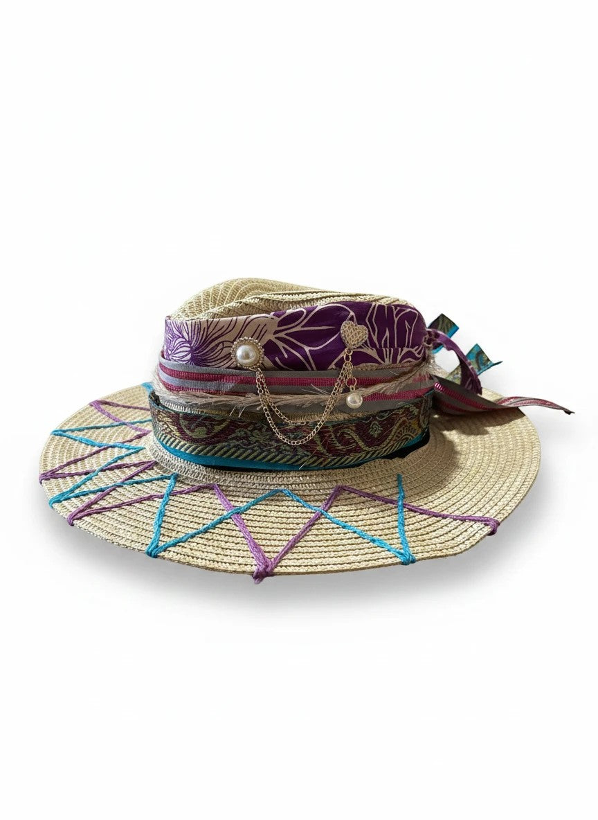 Adams + Hegwood decorative woven straw sun hat with wide brim, purple floral ribbon, pearl & gold charms, and teal/pink geometric stitching.