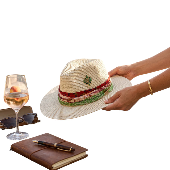 Natalie Adams Hegwood Studio straw fedora with a colorful fabric scarf band and jewel brooch held by hands.
